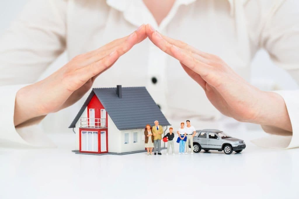 Hände formen ein Dach über einem Modellhaus mit Familie und Auto